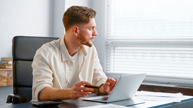 Ein junger Mann hält seine Kreditkarte in der Hand während er aus dem Fenster schaut. Vor ihm steht ein Laptop auf dem Tisch.