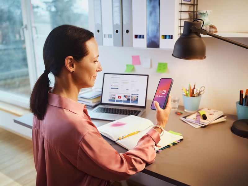 Eine Frau sitzt am Schreibtisch und arbeitet mit Laptop und Mobiltelefon, das sie in der Hand hält. Auf diesem ist die ID Austria zu sehen, die sie für die Anmeldung in den eServices der österreichischen Verwaltung verwendet.