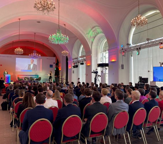 In der Orangerie wurde der Digital Skills Summit abgehalten. Vorne ist die Bühne mit einer großen Rückwand, auf der Staatssekretär Florian Tursky seine Ansprache hält. Die Moderatorin steht am Rande der Bühne bei einem Stehpult. Im Raum sieht man die Gäste - alle Stühle sind besetzt. Die Regie wurde am rechten Rand beim Fenster positioniert.